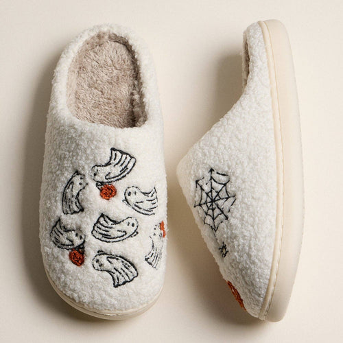 A pair of white sherpa slip-on slippers with embroidered bird and spider motifs, shown on a warm beige background.