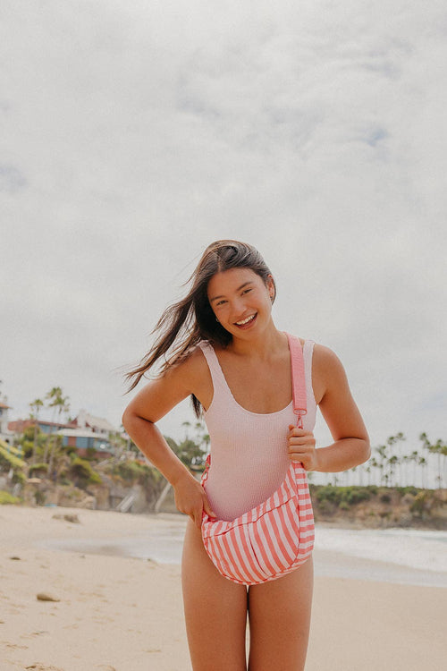 A woman standing on a sandy beach wearing a light pink one-piece swimsuit and carrying a pink and white striped shoulder bag.