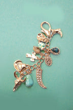 A rose gold charm bracelet featuring multiple religious and cultural symbols, including a cross, Om symbol, crescent moon, skull, and turquoise beads, displayed on a solid teal background.