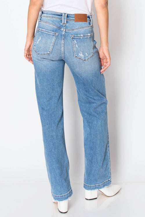 Rear view of a person wearing light blue straight-leg jeans with a brown leather patch at the back waistband, paired with a white top and white pointed-toe heels against a light gray studio background.