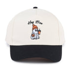An off-white and black baseball cap featuring an embroidered cartoon ghost wearing an orange beanie and holding a pumpkin and an iced coffee, with the text 'Hey Boo.' above it.