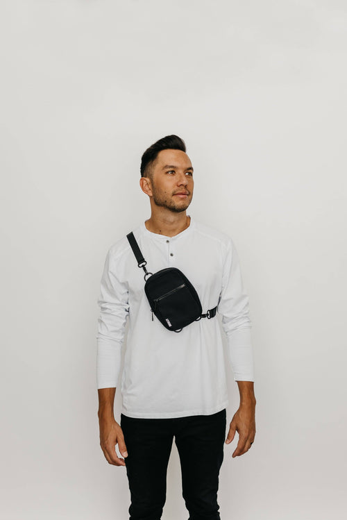 Man wearing a white long-sleeve shirt and black pants with a black crossbody bag on a light gray background
