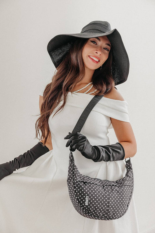 A woman wearing a large black sun hat, black gloves, and a white off-the-shoulder dress carries a black and white polka-dot shoulder bag against a light gray studio background.