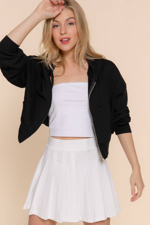 A woman modeling a black cropped zip-up hoodie over a white strapless crop top and a white pleated tennis skirt, posed against a solid beige background.