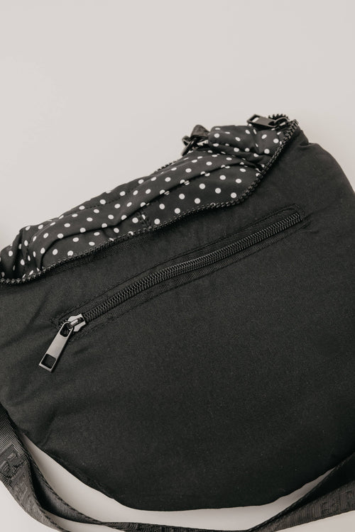 A close-up of a black sling bag with a black and white polka dot patterned fabric lining or trim, featuring a front zippered pocket and a gray patterned strap.