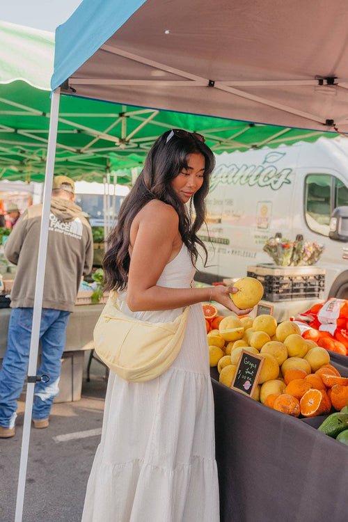 A woman in a white tiered maxi dress stands at an outdoor farmers market under a canopy, holding a yellow fruit next to a display of citrus fruits and wearing a pale yellow crescent shoulder bag.