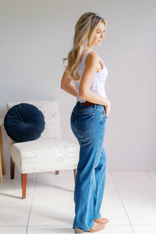 Woman wearing blue jeans and a white top standing in a room with a chair and pillow.