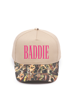 A beige baseball cap with 'BADDIE' printed in bright pink letters on the front, featuring a brown and green camouflage pattern visor.