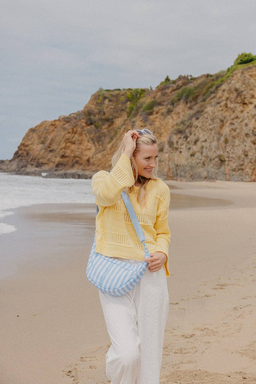 A woman stands on a sandy beach wearing a yellow knit sweater, white pants, and a blue-and-white striped shoulder bag, with rocky cliffs and ocean in the background.