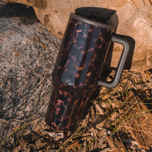 A dark brown and black tortoiseshell-patterned insulated travel mug with a gray handle, resting outdoors on a log and gravel surface.