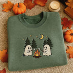 A folded sage green crewneck sweatshirt with a cute Halloween graphic of two ghosts roasting marshmallows by a campfire under a crescent moon, styled on a beige carpet with autumn leaves, pumpkins, and a candle.