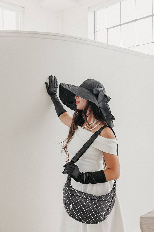 A woman wearing a white off-the-shoulder dress, black wide-brim hat, black gloves, and a black and white polka dot shoulder bag, posing against a white wall in a bright interior.