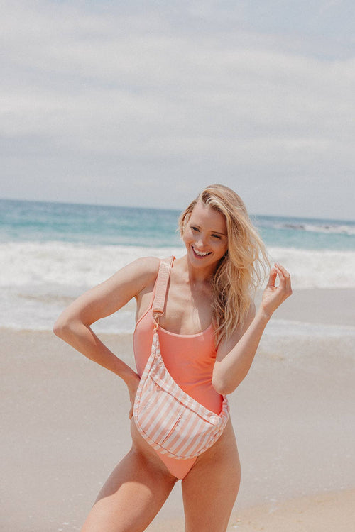 A woman posing on a sandy beach wearing a coral pink one-piece swimsuit and carrying a pink and white striped shoulder bag.