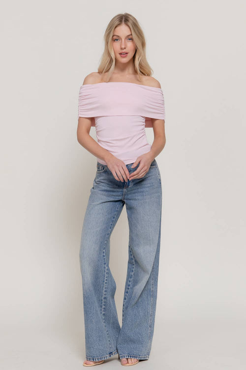 A woman modeling an off-the-shoulder light pink top and light-wash wide-leg blue jeans against a plain light gray studio background.