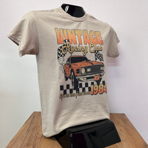 Beige t-shirt with vintage racing car design on a white surface with wooden edge.