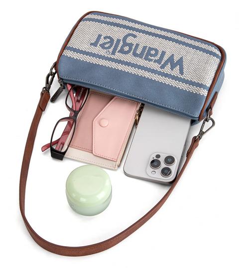 A Wrangler crossbody bag in blue and cream woven fabric with brown leather trim, shown open with accessories inside on a white background. The bag has a blue and cream striped pattern with the Wrangler logo across the top, and is styled with eyeglasses, a pink card holder, a silver smartphone, and a small mint green round container.