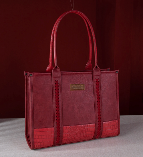 A red leather tote bag with textured panels, crocodile-embossed trim, and a gold-tone brand plaque, photographed against a dark red studio backdrop.