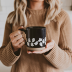 A person in a beige ribbed sweater holds a black ceramic mug with a white ghost pattern around the lower half, photographed against a softly blurred indoor background.