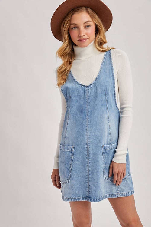Woman wearing a denim dress over a white turtleneck with a brown hat on a light gray background