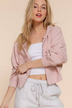 Woman wearing a pink hoodie, white top, and gray pants against a beige background