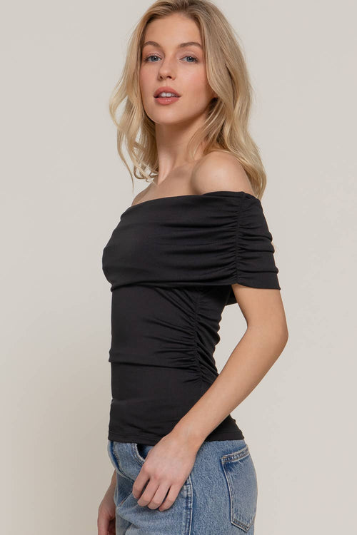 A woman modeling a black off-the-shoulder ruched short-sleeve top paired with light blue jeans, set against a plain light beige studio background.