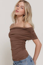 A woman wearing a ruched off-the-shoulder brown top and blue jeans, posing against a light gray studio background.