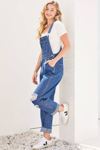 Woman wearing blue denim overalls with a white shirt underneath, standing against a white wall.