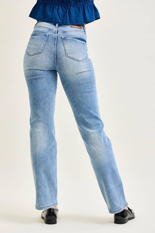 Rear view of a person wearing light blue high-waisted straight-leg jeans, paired with a dark blue ruffled top and black shoes, against a plain light background.
