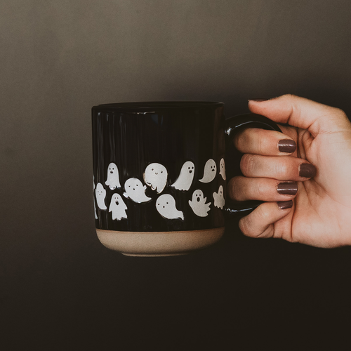 Black mug with ghost pattern held by a hand against a dark background
