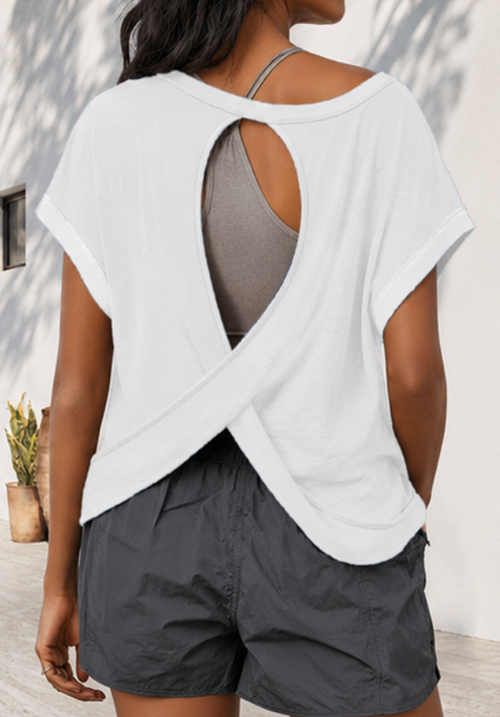 Person wearing a white open-back top over a gray tank top and shorts, standing against a light-colored wall.
