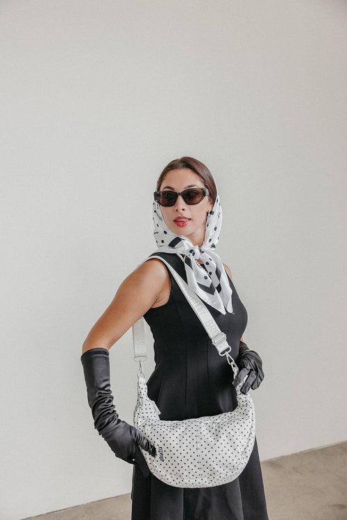 A woman wearing a black sleeveless dress, black gloves, and a black-and-white polka dot headscarf carries a black-and-white polka dot shoulder bag. She is wearing dark sunglasses and posing against a plain light gray wall.