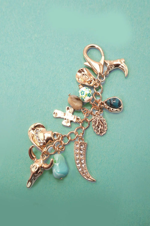 A rose gold charm bracelet featuring multiple religious and cultural symbols, including a cross, Om symbol, crescent moon, skull, and turquoise beads, displayed on a solid teal background.