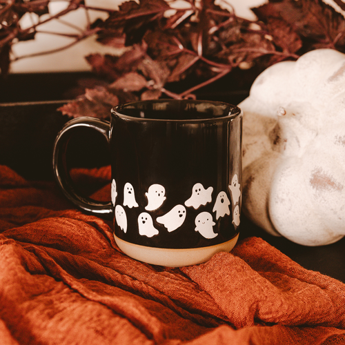 Black mug with ghost design on a red fabric background with pumpkins and leaves.