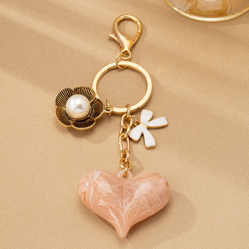 Gold keychain with heart-shaped charm, pearl, and flower on a beige background