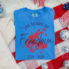 Blue t-shirt with '250 Years of Freedom 1776-2026' text on a red and white patterned surface.