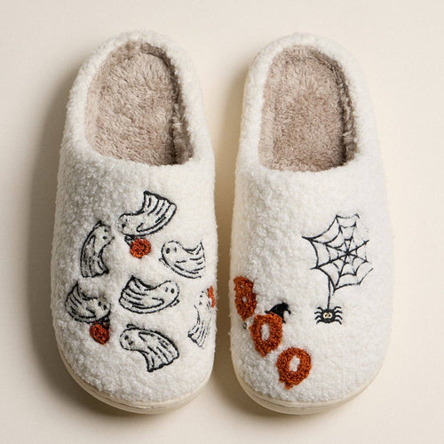 A pair of white sherpa slip-on slippers with embroidered Halloween motifs, including bats, ghosts, a spiderweb, a spider, and orange 'boo' letters, shown on a light beige background.