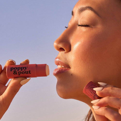 Person applying a red lip balm labeled 'poppy & pout' against a clear blue sky.