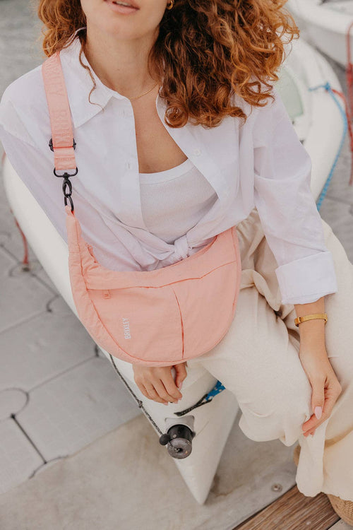A woman with curly red hair wearing a white shirt and cream pants, carrying a light pink crescent-shaped crossbody bag with a black strap and hardware while sitting on a white boat.