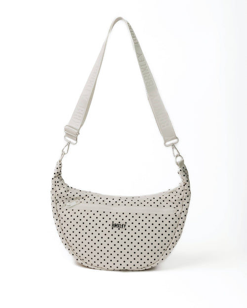 A small crescent-shaped shoulder bag with a black polka dot pattern on a light beige fabric, featuring a matching adjustable shoulder strap and a small black logo on the front.