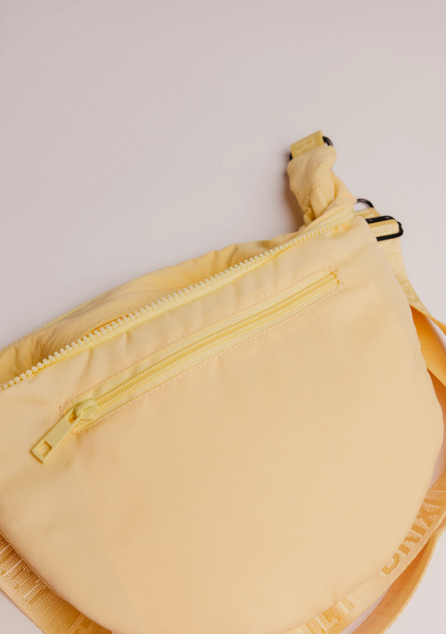 A pale yellow crescent-shaped crossbody bag with a matching yellow zipper and strap, shown on a light off-white background.