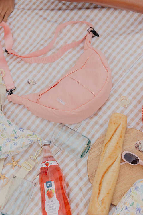 A light pink sling backpack with a wide adjustable shoulder strap, laid out on a white and beige gingham picnic blanket alongside a glass bottle, a baguette, sunglasses, and a straw hat.