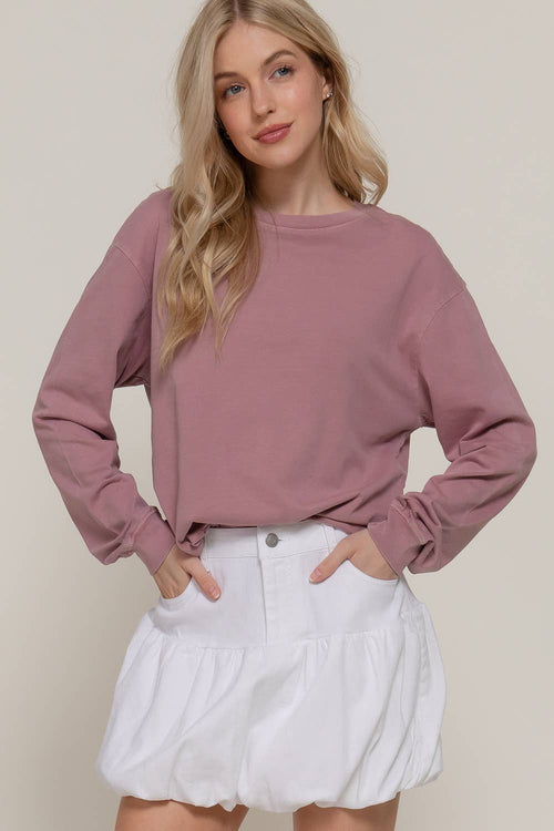 A woman modeling a dusty rose long-sleeve crewneck sweatshirt paired with white ruffled shorts against a solid light beige background.