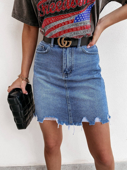 Person wearing a denim skirt with a black belt and holding a black clutch against a white background