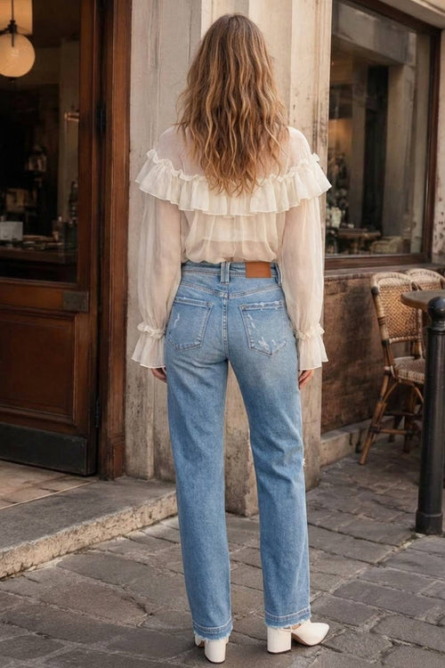 A woman viewed from behind wearing a cream ruffled long-sleeve blouse and light blue straight-leg jeans, standing on a cobblestone street in front of a rustic shop.