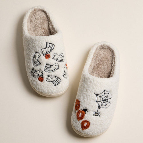 A pair of white boucle slip-on slippers with Halloween-themed embroidery, including ghosts, a spiderweb, a spider, and orange pumpkins, shown on a light beige background.