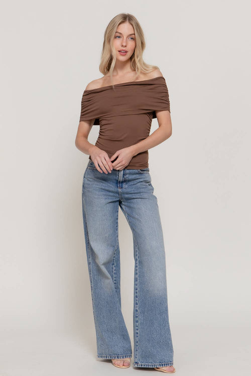 A woman modeling an off-the-shoulder brown ruched top and wide-leg light blue jeans against a plain light gray studio background.