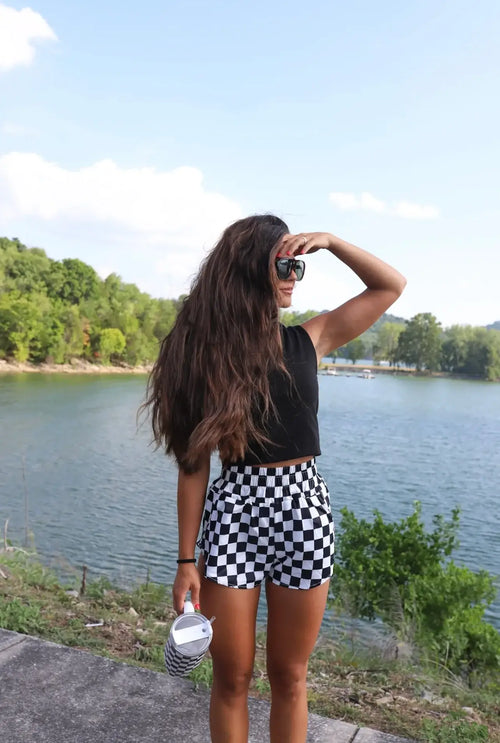 Woman in black top and checkered shorts standing by a lake