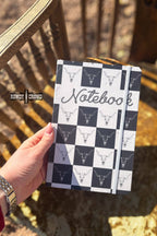 Notebook with checkered pattern and bull head design held by a hand outdoors.