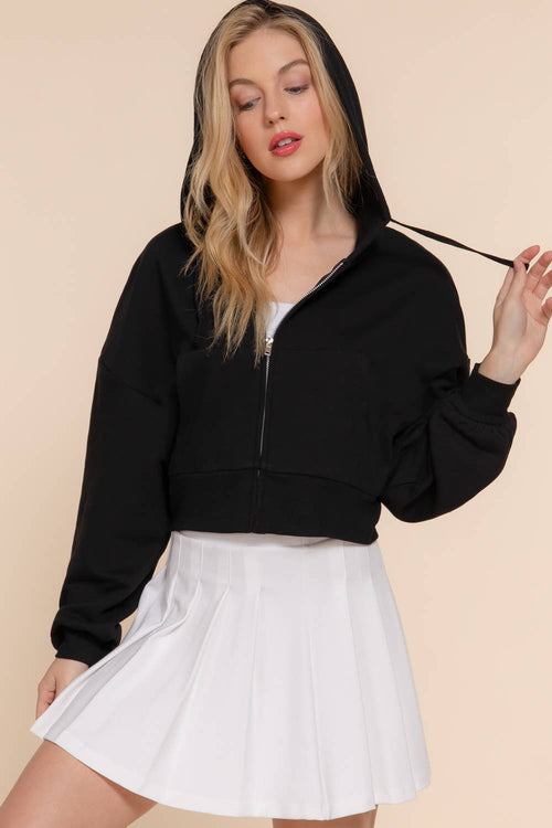 A woman modeling a black cropped zip-up hoodie with a white pleated mini skirt, posed against a solid light beige background.