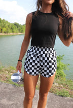 Person wearing black tank top and checkered shorts standing by a lake.